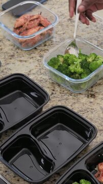 Vertical video: Arm scooping broccoli adding chicken on kitchen counter with empty trays and spoon