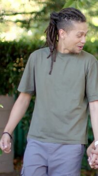 Vertical video: Walking man with locs holding red toy, laughing at call in park, wearing olive tee
