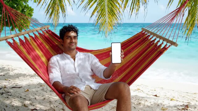 Smiling young man sitting on a red hammock at tropical beach holding blank white smartphone screen, concept for mobile app marketing, travel booking and digital nomad lifestyle