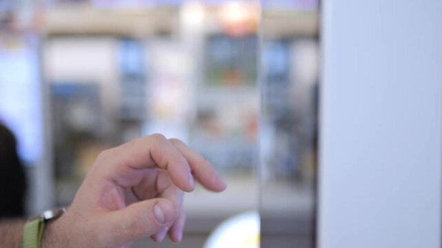 Hand taps touchscreen with focus on gesture. Background blurred, showing indoor setting. Person wears green wristband, arm hair visible. Action suggests interaction with digital device