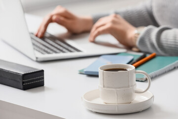 Fototapeta premium Young woman working with laptop at table in office, focus on cup of coffee