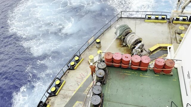 seafarer with water hose washing mooring ropes at the winch and aft mooring station of ship. drums for collect garbage in view. ship proceeding in ocean.