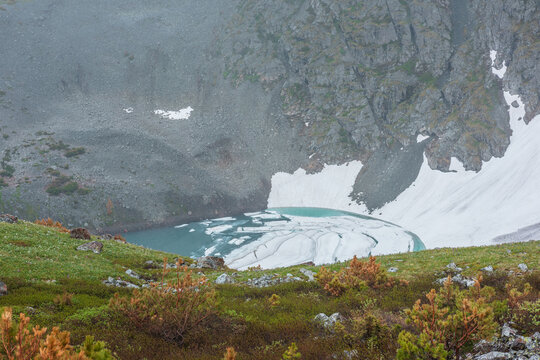 Atmospheric top view from green grassy slope to frozen lake among rocks and snows in mountain cirque in rain. Awesome scenery with ice floes float on turquoise water of alpine lake in rainy weather