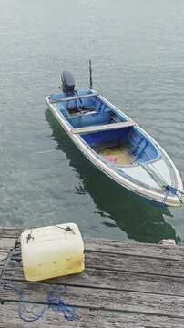 a traditional blue and white fiberglass motorboat gently swaying on calm sea water. The boat is moored next to a rustic wooden dock with a yellow fuel container and blue rope in the foreground
