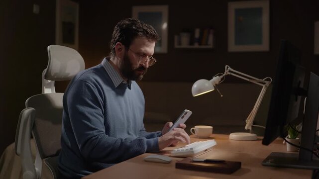 Working from home, freelancer at work concept. Focused man in home office using phone and computer for planning. Camera slowly zooms in