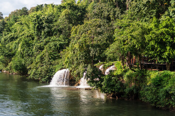 Fototapeta premium A waterfall is flowing into a river, surrounded by lush green trees