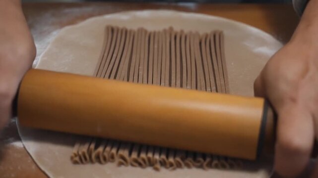 Close-up of hands dynamically rolling a wooden pin across thin, brown, freshly cut noodles, resting on a light dough sheet