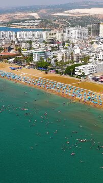 Vertical video. Kizkalesi, Mersin, Turkey. Aerial view of a vibrant city beach with rows of blue sun umbrellas, crowded shoreline and people swimming in turquoise Mediterranean water under clear summ