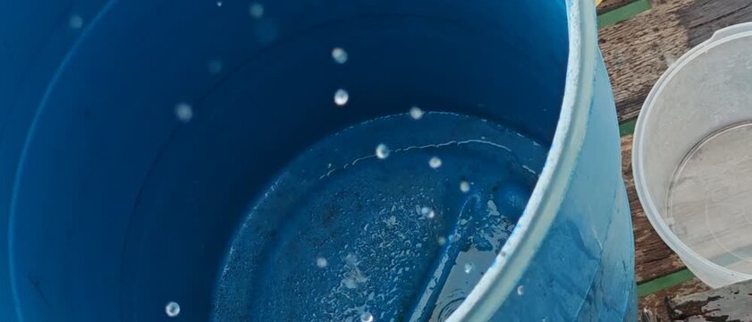A close-up, high-angle shot of clear water droplets pouring and splashing into a large blue plastic storage barrel