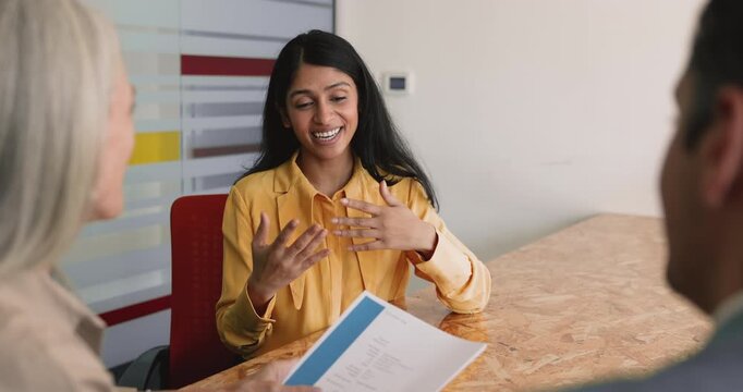 Positive confident young Indian job candidate telling about professional skills to recruiters, employers, speaking at meeting table, explaining paper resume on interview meeting, smiling