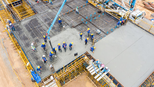 Construction Site Concrete Pour: An aerial view showcases a bustling construction site where workers in hard hats are actively pouring concrete, creating a solid foundation for a new structure.