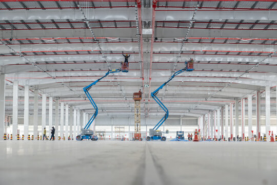 Construction of Large Warehouse Interior: Two aerial work platforms are positioned inside a massive, unfinished warehouse, with workers visible carrying out construction and installation tasks.