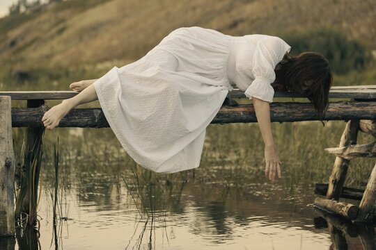 Barefoot woman in a vintage white dress lies on a small wooden bridge over a river and reaching out to the water, magic atmosphere.