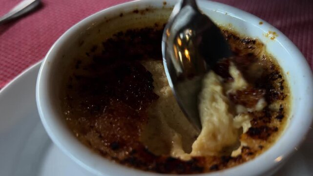 Close view of spoon breaking caramelized sugar crust on creme brulee dessert served in ceramic ramekin at restaurant table.