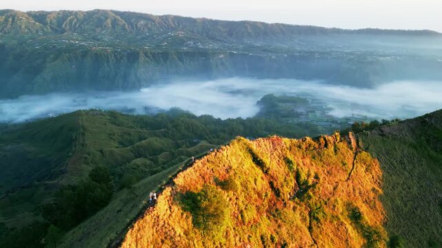 Golden sunrise with hikers walking along rugged volcanic ridge above mist filled caldera valley and dramatic tropical mountain landscape in Bali aerial drone footage.