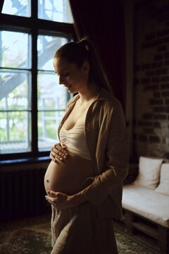 A pregnant woman stands in a warmly lit living room, softly cradling her belly with a peaceful expression. The room features large windows that allow natural light, creating a serene atmosphere