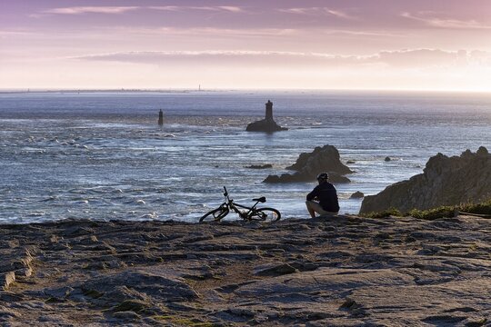 Cyclist sitting on rocky coast, taking a break, Pointe du Raz, Phare de la Vieille, silhouette against evening sky, Cap Sizun, D&eacute;partement Finist&egrave;re, Bretagne, France