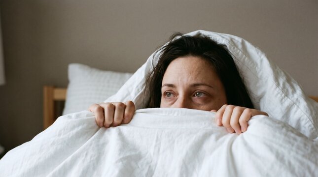 Woman Hiding Under Blanket with Fearful Expression in Bedroom.