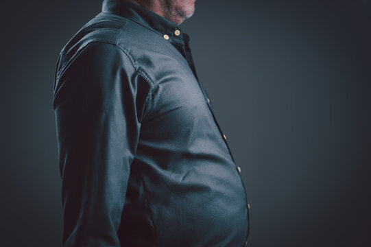Mature man wearing dark buttoned shirt stands in profile against gray background highlighting midsection weight gain as part lifestyle health assessment regarding body mass index physical fitness.