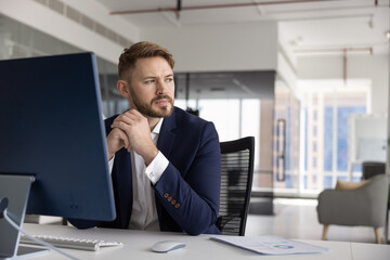 Fototapeta premium Young businessman in suit sits at his office desk, looking away with thoughtful expression, deep in contemplation, planning strategy, making decision, thinks on future opportunities. Business vision