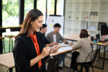 Fototapeta premium Young Asian Businesswoman Using Smartphone in Modern Office with Team Working in Background