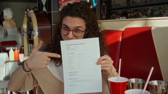 Medium portrait of nerdy gen Z Caucasian male student with curly long hair, eyeglasses covering face with exam grade sheet, lowering and pointing at successful score, while celebrating at burger diner