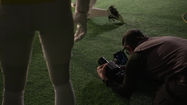 Medium backstage shot of male cinematographer lying on ground, recording moment of kickoff for American football show or TV commercial from low angle on professional video camera
