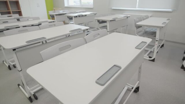 Empty school desks stand in modern light classroom. Soft morning sun creates calm study atmosphere. Organized classroom awaits students