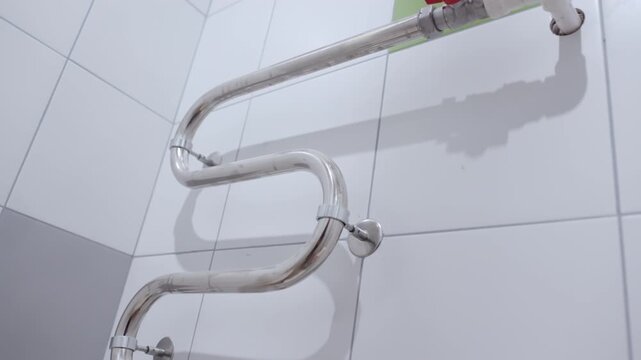 Curved metal radiator pipe features red valves in clean tiled bathroom. Rubber gloves dry on metallic surface creating sterile environment