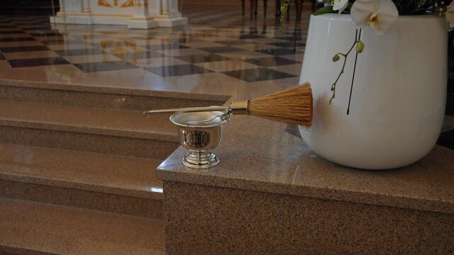 Detailed close-up shot of a holy water sprinkler and silver vessel on polished marble steps. Ornate white planter with flowers, checkered floor, and soft natural light create a sacred mood.