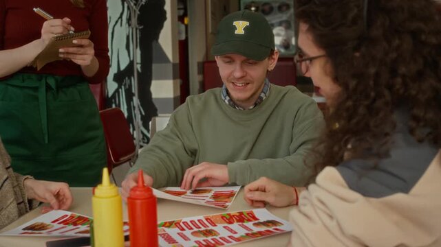Medium close-up of hungry young college students talking to waitress taking order at burger restaurant, asking for biggest portions, gesticulating and laughing while enjoying hanging out together