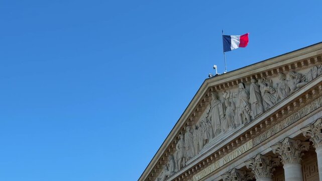 Tricolore flag waving in the wind atop the pediment of the Palais Bourbon, home of the French National Assembly in Paris, against a clear blue sky on a sunny day in the capital of France