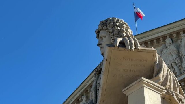 Monumental sculpture of Chancellor d'Aguesseau on the facade of the Assembl&eacute;e Nationale in Paris, with the French flag waving against a clear blue sky, symbolizing law and French heritage