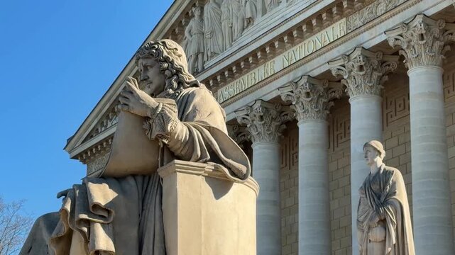 Detailed view of the classical facade of the Assembl&eacute;e Nationale in Paris, France, showcasing its intricate statues and Corinthian columns under a clear blue sky on a sunny day