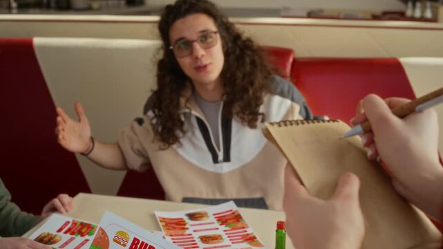 Medium close-up of hands of unrecognizable waitress taking order from cheerful group of zoomers sitting at table at burger cafe, writing down on notepad, providing hospitality service