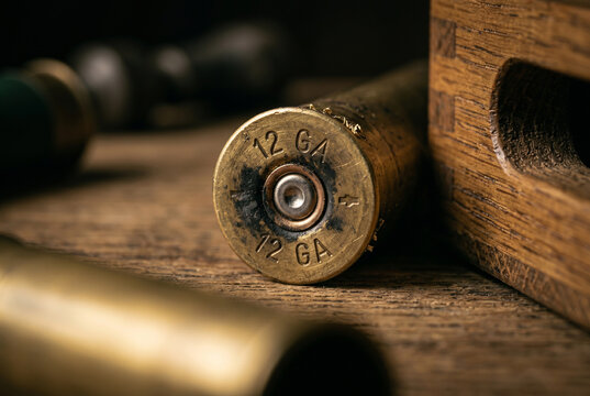 Macro close-up of a tarnished 12-gauge brass shotgun shell headstamp, showing the indented primer pocket surrounded by dark black powder soot on an ash wood workbench surface.