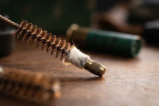 Macro close-up of a twisted bronze wire bore brush with a cotton patch and knurled brass tip on a wooden workbench, with a green shotgun shell blurred in the moody background.