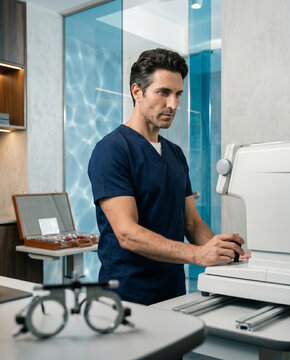 Medium shot of a doctor operating a diagnostic workstation with an integrated joystick. Sleek white structural body, anodized tracks, open leather lens case, and refracted glass lights.
