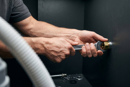 Macro action focuses on a plumber's tensed forearms and hands, gripping a steel wrench to tighten a brass nut on copper pipe threads in an anthracite cabinet with mineral porcelain backdrop.