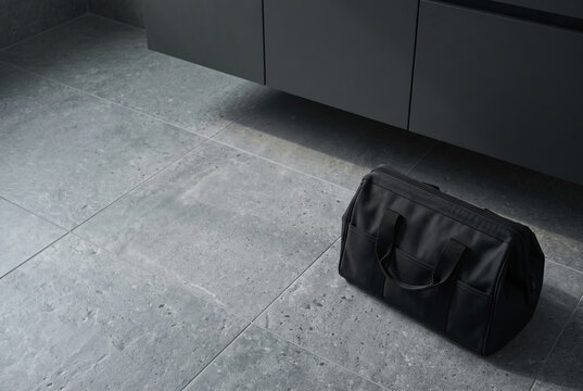 Wide horizontal still life shot from a slight high-angle captures a premium, unbranded, unzipped high-density black nylon tool bag fully packed on a large-format grey stone porcelain bathroom tile.