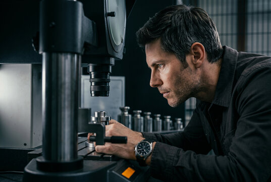 A senior metrology specialist inspects a micro-machined steel component using a high-precision optical comparator microscope in a state-of-the-art industrial testing laboratory.
