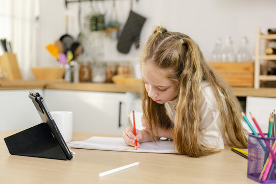 A young girl does homework at a kitchen table, writing in a notebook while using a tablet for study. back to school concept