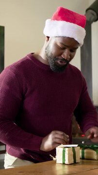 Vertical video: Picking parcel, man in sweater and Santa hat tying ribbon for holiday gifts at home