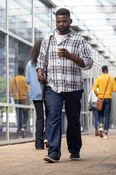 Adult African man walking along glass walkway plaid-shirt holding to-go coffee and messenger bag