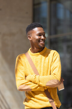 African American man smiling while holding coffee and crossbody bag by building in mustard sweater
