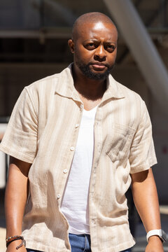 African man walking under metal beams at transit hub wearing beige shirt, smartwatch, bracelets