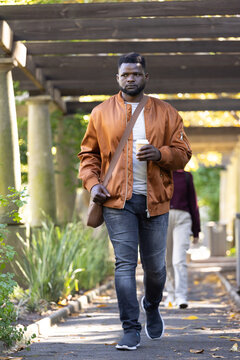 African American adult man walking under pergola toward camera wearing bomber jacket holding cup