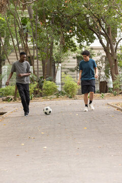 Diverse adult male friends in athletic clothing walking and kicking soccer ball on paved driveway