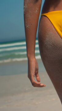 Vertical video: Walking woman in yellow bikini bottom along shore; sand on skin, copy space