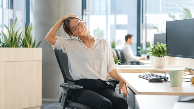 Office worker woman stretching her neck at a desk in a bright modern open-plan workspace
